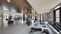 This single-level fitness center is designed to support a comprehensive workout experience, combining thoughtful architectural details with a wide array of professional-grade equipment. The space is divided into distinct functional zones, each tailored to different aspects of physical training.

The main workout area features a striking wood-paneled ceiling that adds warmth and visual interest, contrasting with the light-toned walls and polished epoxy flooring below. Ceiling fans are strategically positioned to maintain airflow throughout. Recessed lighting supplements the abundant natural light that streams in through large, floor-to-ceiling windows and paneled glass doors, offering views of an outdoor pool area and surrounding grounds.

Strength training is well-represented with a multi-station cable machine, free-weight benches, a full dumbbell rack, a heavy punching bag, and various resistance machines. A dedicated cardio zone houses treadmills, an elliptical, and a stair climber, all positioned near mirrored walls that enhance the sense of space. Wall-mounted televisions are installed throughout for entertainment during workouts. A plyo box and additional functional training accessories round out the facility's offerings.