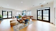 This 580 square foot living space showcases a modern and inviting atmosphere. The open floor plan maximizes the use of space, creating a seamless flow between different functional areas. Large glass doors and windows flood the room with natural light, offering views of the exterior and creating a connection with the outdoors.

The space features warm wood flooring throughout, adding a touch of natural elegance. The furnishings are carefully chosen to blend comfort with style, including a tan leather sofa, blue accent chairs, and woven pouf ottomans. A round dining table with modern chairs provides a dedicated eating area.

A striking blue accent wall adds a pop of color, contrasting beautifully with the predominantly white walls. The room is adorned with tasteful decorations, including framed artwork and potted plants, which contribute to a fresh and lively ambiance.

The space also includes practical amenities such as a wall-mounted TV, a compact kitchenette area, and what appears to be a game table, possibly for shuffleboard. Recessed lighting in the ceiling provides even illumination throughout the space.

This versatile living area seems designed to accommodate various activities, from casual lounging to entertaining guests, making it an ideal space for modern, multifunctional living.