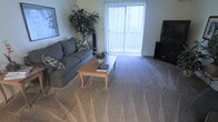 This single-floor living space centers around a well-appointed living room designed for both comfort and functionality. The room is finished with wall-to-wall carpeting in a neutral tone, contributing to a warm and inviting atmosphere throughout. A large sliding glass door dominates one wall, fitted with vertical blinds that allow for adjustable natural light and provide access to an outdoor area. The generous flow of daylight enhances the overall brightness of the space.

The living room is arranged to maximize usability, featuring a spacious grey sofa accented with decorative pillows and positioned to face a television setup housed on a dark media console. A wooden coffee table serves as the centerpiece of the seating area, topped with a small potted plant and reading material, adding a lived-in, homey quality. A lush indoor tree positioned near the sliding door adds a touch of greenery and visual interest. A framed piece of wall art is mounted near the entrance, providing a subtle decorative accent. The overall ambiance is relaxed and comfortable, making it a practical everyday living environment.