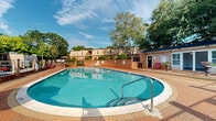 Located at 702 Edwards Rd in Greenville, South Carolina, this community space centers around a generously sized oval swimming pool that serves as the focal point of the outdoor area. The pool is enclosed by a classic brick wall perimeter, lending the space a sense of defined structure and privacy while mature, leafy trees rise above the boundary, creating a natural canopy that adds shade and a lush, park-like atmosphere.

The pool deck is finished with attractive brick paving that surrounds the water on all sides, providing ample room for lounging and socializing. White lounge chairs and sun beds are positioned along the poolside, offering comfortable spots to relax, while a patio table with a market umbrella and additional seating create a dedicated outdoor dining and gathering area. Safety equipment, including a life ring, is visibly mounted, and posted signage indicates standard pool rules.

A small utility structure is accessible from the deck, likely serving as a pool equipment or storage room. The residential buildings visible beyond the perimeter feature balconies and a mix of brick and siding exteriors. The interior image reveals a bright, clean room with light speckled flooring, white walls, and ceiling fans, reflecting a practical and well-maintained single-floor layout. Overall, the space offers a welcoming and functional outdoor recreational environment suited for community use.