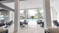 This expansive outdoor amenity space at Inspira Lifestyle Living is designed with a resort-inspired aesthetic, blending open-air relaxation with functional gathering areas. The covered pavilion serves as the centerpiece, featuring a crisp white ceiling with recessed lighting, dark exposed beam accents, and ceiling fans to ensure comfort in warmer conditions. The flooring throughout is a light-toned stone tile that extends seamlessly across all zones, creating visual continuity.

The lounge area is furnished with multiple dark-framed sectional sofas and chairs upholstered in white, arranged symmetrically to encourage social interaction. A live-edge wood coffee table anchors one seating cluster near the entrance, while a wall-mounted television and fireplace provide additional comfort. French doors lead into an interior clubhouse, offering a smooth transition between indoor and outdoor spaces.

Adjacent to the lounge is a recreational zone featuring a pool table, bar-height seating, and additional dining tables with rattan chairs. Beyond the covered structure, a large resort-style swimming pool is framed by palm trees and lounge chairs, with multi-story residential buildings visible in the background. The overall ambiance is bright, modern, and tropical, making this an ideal space for both relaxation and socializing.