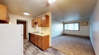 This single-floor unit offers a practical and comfortable layout designed for everyday living. The kitchen is equipped with warm-toned wood cabinetry providing ample storage, a countertop sink, and space for essential appliances including a refrigerator and dishwasher. The kitchen flows naturally into the adjacent living area, creating an open, connected feel throughout the main space. Underfoot, the kitchen features durable wood-look plank flooring, while the living area transitions to wall-to-wall carpeting for added comfort. A large window in the living area fills the space with natural light and offers a view of the surrounding greenery outside. A ceiling fan overhead provides air circulation throughout the open area. A textured ceiling adds a classic residential character to the space. A baseboard heater along the living room wall ensures warmth during cooler months. A doorway visible from the main area provides access to additional rooms within the unit, suggesting a functional and well-organized floor plan. Overall, the space presents a clean, straightforward layout suited for comfortable, practical living.