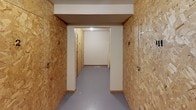 This single-floor storage facility is organized around a central corridor that provides access to a series of individually numbered storage lockers lining both sides of the hallway. The lockers are constructed from oriented strand board (OSB) panels, giving the space a utilitarian, no-frills aesthetic. Each unit is secured with its own latch hardware and is clearly numbered for easy identification, with unit numbers visible throughout the corridor.

The flooring throughout consists of a smooth, painted concrete surface in a neutral gray tone, providing a durable and easy-to-maintain base. The ceilings feature a textured finish and are equipped with overhead lighting fixtures that illuminate the corridor and locker areas.

Access to the storage area is controlled through a solid wood interior door fitted with a door closer mechanism and a posted notice indicating that ownership and management assume no responsibility for stolen, lost, or damaged items left in the storage lockers. The corridor branches into additional sections, expanding the capacity of the facility and accommodating a greater number of storage units. The overall layout is straightforward and functional, designed to maximize storage density while maintaining clear walkway access to each individual locker.