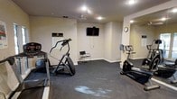 This fitness center provides a dedicated space for exercise, designed with functionality in mind. The room features neutral-toned walls with white crown molding trim and dark carpeted flooring that helps define the workout area. Recessed lighting and a central ceiling fixture ensure the space is well-lit throughout.

The cardio equipment selection includes an elliptical trainer, a stationary bike, and an additional cardio machine, giving users several options for aerobic exercise. A wall-mounted flat-screen television is positioned for easy viewing during workouts, adding entertainment value to training sessions. A wall clock allows users to keep track of time conveniently.

A full-length mirror on one wall visually expands the room while serving a practical purpose for monitoring form. A simple chair provides a resting spot, and a water cooler ensures hydration is readily accessible. A posted fitness center rules and guidelines sign indicates this is a shared facility. The overall layout is clean and organized, maximizing the usable floor space for movement and equipment placement.