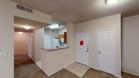 This single-story space offers a functional and straightforward layout designed for comfortable everyday living. The entryway opens to a tiled foyer flanked by two white panel doors, providing a welcoming transition into the main living areas. A half-wall breakfast bar separates the kitchen from the living space, creating an open-concept feel while maintaining defined areas. The kitchen is well-equipped, featuring warm medium-toned wood cabinetry extending to the ceiling for ample storage, granite-style laminate countertops, a white electric range with coil burners, an over-the-range microwave, a refrigerator, and a double stainless steel sink. Tile flooring runs throughout the kitchen and entryway, while neutral-toned carpet extends across the living room and private rooms. Each bedroom features a ceiling fan with integrated lighting and windows fitted with horizontal blinds, allowing natural light to filter in. The bathroom is outfitted with a tiled tub-surround accented by a decorative horizontal band, stainless steel grab bars for accessibility, a vanity with a stone-style countertop, and a large mirror. The overall color palette is warm and neutral throughout, lending a cohesive and understated ambiance to the entire space.