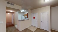 This single-story space offers a functional and straightforward layout designed for comfortable everyday living. The entryway opens to a tiled foyer flanked by two white panel doors, providing a welcoming transition into the main living areas. A half-wall breakfast bar separates the kitchen from the living space, creating an open-concept feel while maintaining defined areas. The kitchen is well-equipped, featuring warm medium-toned wood cabinetry extending to the ceiling for ample storage, granite-style laminate countertops, a white electric range with coil burners, an over-the-range microwave, a refrigerator, and a double stainless steel sink. Tile flooring runs throughout the kitchen and entryway, while neutral-toned carpet extends across the living room and private rooms. Each bedroom features a ceiling fan with integrated lighting and windows fitted with horizontal blinds, allowing natural light to filter in. The bathroom is outfitted with a tiled tub-surround accented by a decorative horizontal band, stainless steel grab bars for accessibility, a vanity with a stone-style countertop, and a large mirror. The overall color palette is warm and neutral throughout, lending a cohesive and understated ambiance to the entire space.