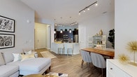 This well-appointed single-floor residence showcases a bright, open-concept layout that effortlessly connects the living, dining, and kitchen areas into one cohesive living space. The kitchen is a standout feature, boasting dark espresso cabinetry that extends to the ceiling with decorative greenery accents, stainless steel appliances including a refrigerator and microwave, a stylish tile backsplash, and a peninsula island with a light countertop and seating for two with modern bar stools. Pendant lighting elegantly illuminates the island, while a track lighting system overhead provides flexible ambient light throughout. The dining area comfortably accommodates a full-sized table and chairs, complemented by a decorative bar shelving unit. The living area is open and airy, anchored by light-toned furnishings and a patterned area rug. Warm wood-look plank flooring runs throughout the entire space, tying each zone together harmoniously. The entry foyer is clearly defined, offering a welcoming transition into the home. White walls and abundant artificial lighting contribute to a clean, contemporary ambiance that feels both spacious and inviting.