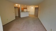 This single-floor unit offers a practical and comfortable layout with carpeted flooring throughout the main living spaces. The living area is generously sized, featuring neutral-toned walls, a textured ceiling, and a large window fitted with vertical blinds that allows natural light to fill the room, creating a bright and airy atmosphere.

Adjacent to the living area, the kitchen is well-equipped and designed for functionality. It features warm wood cabinetry, a white refrigerator, recessed lighting, and a breakfast bar peninsula with a dark countertop that separates the kitchen from the living space. This open-concept design promotes a seamless flow between cooking and entertaining areas.

A ceiling fan is centrally positioned in the living space, providing efficient air circulation. Multiple doorways visible from the main area lead to additional rooms, suggesting a practical floor plan with accessible private spaces. The overall design emphasizes clean lines and neutral tones, creating a versatile interior that can accommodate a variety of furnishing styles.