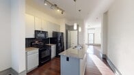 Located at 7105 NC Highway 86 in Chapel Hill, NC, this two-floor residence offers a thoughtfully designed interior with a range of quality finishes. The kitchen is a standout feature, equipped with a center island topped with granite countertops and an integrated sink, white cabinetry, a black tile backsplash, and black appliances including a gas range, built-in microwave, and French door refrigerator. Pendant lighting and a track light fixture illuminate the space, which flows seamlessly into a bright, open living area beyond. Rich, dark hardwood flooring runs throughout the main living spaces, complementing the clean white walls and gray-accented trim. The living area is generously proportioned, with large windows and a glass-paneled door that invites abundant natural light. Two bathrooms serve the home: one features a granite vanity, large-format tile flooring, and a glass-enclosed shower with tiled surround, while the second offers a vessel sink, granite countertop, and a full tub with a dark tile surround. An additional room on the upper floor is finished with plush carpet and a ceiling fan, providing a comfortable and quiet retreat. Throughout the home, consistent use of gray trim, neutral tones, and quality materials creates a cohesive, modern aesthetic.