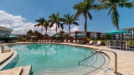 Located at Sierra Grande Apartments in Naples, Florida, this single-story amenity complex is designed to deliver a resort-style experience in a lush, tropical setting. At the heart of the space is a generously sized, curved swimming pool with crystal-clear turquoise water, accessible via wide entry steps and surrounded by an expansive tiled pool deck. Rows of lounge chairs and teal-accented umbrellas line the deck, offering ample space for sunbathing and relaxation beneath the Florida sun. Towering palm trees frame the area, providing natural shade and a distinctly tropical ambiance.

Adjacent to the pool, a covered outdoor pavilion serves as a versatile gathering and recreation area. Supported by arched columns, the pavilion features a vaulted white ceiling with dual ceiling fans to ensure comfortable airflow. A ping pong table and the Sierra Grande Apartments branded mat anchor the space, making it ideal for casual recreation. The pavilion connects directly to an interior clubhouse space through large sliding glass doors, creating a seamless indoor-outdoor flow.

The clubhouse building itself showcases Mediterranean-inspired architecture, characterized by a terracotta tile roof, arched entryways, stucco exterior, and decorative lantern-style lighting. The entire complex is thoughtfully landscaped, creating a welcoming and visually rich community amenity space.