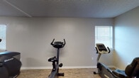This single-floor fitness space at Westtowne Cottages is purpose-built for exercise and wellness. The room is outfitted with a comprehensive selection of cardio equipment, including a treadmill, an elliptical trainer, an upright stationary bike, and a recumbent bike, providing a well-rounded workout environment suitable for a range of fitness levels and preferences.

The room features durable light wood-tone laminate flooring, which is both practical and easy to maintain in a high-use fitness setting. The walls are finished in a neutral gray tone, lending the space a clean, focused atmosphere. A textured ceiling adds a subtle design element overhead, while flush-mounted lighting ensures the room is well lit throughout. A window fitted with horizontal blinds allows for natural light while maintaining privacy, contributing to a bright and energizing workout environment.

A doorway connects directly to an adjacent full bathroom, complete with a vanity, mirror, and sink — offering added convenience for post-workout routines. A hallway is also accessible from the fitness room, providing easy flow throughout the broader layout. The overall design is functional and straightforward, making this a highly practical and well-equipped space for daily fitness use.