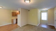 This single-floor space showcases a practical open-concept layout that seamlessly connects the living area to the kitchen. The living area is finished with wall-to-wall carpeting, providing a comfortable underfoot feel, while the kitchen transitions to hard surface flooring for easy maintenance. Warm beige walls run throughout, creating a neutral and cohesive atmosphere.

The kitchen is equipped with wood-toned cabinetry, a white dishwasher, and a sink positioned along the counter, offering efficient functionality within a compact footprint. A half-wall partition separates the kitchen from the living space, maintaining an open feel while providing a subtle definition between areas.

The textured popcorn ceiling features a central flush-mount light fixture, providing ambient illumination throughout the main living space. HVAC vents are strategically positioned in the ceiling and walls to ensure consistent climate control. A window along the far wall allows natural light to filter into the space, brightening the interior. White baseboards frame the perimeter of the rooms, adding a clean, finished look to the overall design.
