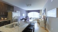This single-floor residence showcases an open-concept layout that seamlessly connects the kitchen, dining, and living areas into one cohesive and functional space. The kitchen is equipped with stainless steel appliances, including a full-size refrigerator, and features a granite-topped island with bar seating for two, complemented by a deep single-basin sink with a modern gooseneck faucet. The granite countertop displays rich, neutral tones that contrast elegantly with the lavender-painted walls found throughout the space.

The dining area sits adjacent to the kitchen island and accommodates a small table with seating for multiple guests, anchored beneath track lighting mounted on the white ceiling. The living room receives generous natural light through a large arched window dressed with a decorative patterned valance and sheer white curtains. The seating area is furnished with neutral-toned chairs and a sofa, arranged around a central coffee table atop an area rug. Light hardwood flooring extends throughout the entire space, enhancing the bright, airy feel. The overall ambiance is warm, modern, and thoughtfully coordinated.