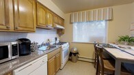 This single-floor space centers around a efficiently designed kitchen that balances functionality with a warm, homey aesthetic. The kitchen is outfitted with raised-panel wood cabinetry in a honey-toned finish, providing ample upper and lower storage. A white gas range sits beneath the upper cabinets, accompanied by an integrated range hood, while a double basin sink with a granite-look countertop offers practical workspace. The white tile backsplash adds a clean, classic contrast to the warm wood tones throughout. Natural light enters through a notably sized window dressed with a striped fabric valance and horizontal blinds, brightening the space and offering a glimpse of outdoor greenery. A baseboard heating unit is positioned beneath the window, ensuring year-round comfort. The flooring features a neutral, low-pile surface that complements the overall palette. A small dining area with wooden bar-height seating is tucked adjacent to the kitchen window, making efficient use of the available space. The overall ambiance is warm and practical, with a traditional design sensibility.