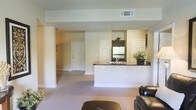 This single-floor residence offers an open-concept layout that connects the living area and kitchen in a cohesive, functional design. The living room is carpeted in a neutral tone, providing a warm and comfortable foundation for the space, and is furnished with classic leather seating. A peninsula-style kitchen counter with a dark granite countertop serves as the visual and functional centerpiece, providing both workspace and a natural dividing element between the kitchen and living areas. The kitchen is equipped with dark cabinetry, stainless steel appliances including a refrigerator, and a built-in sink along the peninsula. Soft, cream-colored walls throughout create a bright and airy ambiance, complemented by recessed overhead ventilation. The entryway is clearly visible from the living area, featuring a classic paneled door that reinforces the clean, traditional aesthetic. Additional doors visible within the space suggest access to further rooms or storage, contributing to the overall practicality of the layout. Decorative touches such as potted greenery add a subtle, natural warmth to the kitchen area. The overall design balances simplicity with functionality, making it an inviting and well-organized living environment.