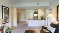 This single-floor residence offers an open-concept layout that connects the living area and kitchen in a cohesive, functional design. The living room is carpeted in a neutral tone, providing a warm and comfortable foundation for the space, and is furnished with classic leather seating. A peninsula-style kitchen counter with a dark granite countertop serves as the visual and functional centerpiece, providing both workspace and a natural dividing element between the kitchen and living areas. The kitchen is equipped with dark cabinetry, stainless steel appliances including a refrigerator, and a built-in sink along the peninsula. Soft, cream-colored walls throughout create a bright and airy ambiance, complemented by recessed overhead ventilation. The entryway is clearly visible from the living area, featuring a classic paneled door that reinforces the clean, traditional aesthetic. Additional doors visible within the space suggest access to further rooms or storage, contributing to the overall practicality of the layout. Decorative touches such as potted greenery add a subtle, natural warmth to the kitchen area. The overall design balances simplicity with functionality, making it an inviting and well-organized living environment.