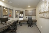 This business center is a compact, efficiently designed workspace that offers residents or occupants a fully functional environment for productivity. The room features an L-shaped, built-in dark wood desk that wraps along two walls, providing ample surface area for multiple workstations equipped with desktop computers and keyboards. Ergonomic task chairs accompany each station, ensuring comfortable use during extended sessions. A multifunction printer/copier is conveniently integrated into the workstation layout, supporting a range of office needs. Above the desk, a row of dark wood wall-mounted cabinets provides additional storage, complementing the warm, neutral tone of the room's walls and carpet flooring. Interior windows with gray-trimmed frames offer a view into the adjacent fitness center, connecting the two amenity spaces visually while maintaining acoustic separation. A glass-paneled sliding door provides easy entry and exit, contributing to the open, accessible feel of the room. Recessed ceiling lighting ensures the space is well-lit throughout. Informational signage near the entrance outlines usage guidelines for the business center, reflecting a thoughtfully organized and professionally maintained amenity space.