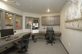 This business center is a compact, efficiently designed workspace that offers residents or occupants a fully functional environment for productivity. The room features an L-shaped, built-in dark wood desk that wraps along two walls, providing ample surface area for multiple workstations equipped with desktop computers and keyboards. Ergonomic task chairs accompany each station, ensuring comfortable use during extended sessions. A multifunction printer/copier is conveniently integrated into the workstation layout, supporting a range of office needs. Above the desk, a row of dark wood wall-mounted cabinets provides additional storage, complementing the warm, neutral tone of the room's walls and carpet flooring. Interior windows with gray-trimmed frames offer a view into the adjacent fitness center, connecting the two amenity spaces visually while maintaining acoustic separation. A glass-paneled sliding door provides easy entry and exit, contributing to the open, accessible feel of the room. Recessed ceiling lighting ensures the space is well-lit throughout. Informational signage near the entrance outlines usage guidelines for the business center, reflecting a thoughtfully organized and professionally maintained amenity space.