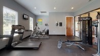 The fitness center at Huntington Park Manor in Nashville, Tennessee is a thoughtfully designed workout facility that provides residents with a comprehensive exercise environment. The space features durable rubber flooring throughout, providing a safe and functional surface ideal for a wide range of physical activities.

The cardio zone is equipped with multiple treadmills, an elliptical trainer, and a stationary bike, each positioned near windows with slatted blinds that allow natural light to filter in. Wall-mounted televisions are conveniently placed to keep residents entertained during workouts. The strength training area includes a multi-function cable machine and an adjustable workout bench, accommodating a variety of resistance exercises.

A dedicated free weights section features a full rack of medicine balls and an assortment of stability and exercise balls in multiple sizes, supporting functional fitness and flexibility training. The walls are painted in a cool gray palette with a bold orange accent wall that adds energy and visual interest to the space.

Large glass panel walls and a glass-paned exterior door connect the fitness center to an adjacent clubhouse area, creating an open, airy feel. Additional windows bring in outdoor views and ample daylight. Recessed lighting provides even illumination throughout. The facility is well-maintained and designed to support a broad range of fitness routines for residents of all experience levels.