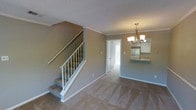 This two-floor residence offers a thoughtfully arranged layout with distinct living and dining areas visible from the main entry point. The living area is finished with wall-to-wall carpet in a neutral beige tone, contributing to a warm and comfortable atmosphere. Soft grey-green walls are complemented by crisp white crown molding and baseboards, adding a clean, polished look throughout.

A staircase with white-painted wooden railings connects the two floors, positioned prominently along one wall of the living area. Overhead, a classic chandelier with candelabra-style lighting serves as a focal point in the dining area, providing both functional illumination and an elegant touch.

An open passageway leads into the kitchen, where white cabinetry and a stainless steel refrigerator are visible, suggesting a practical and well-appointed cooking space. A door at the end of the hallway opens toward what appears to be a bright area with natural light filtering through a window.

The textured ceiling adds depth to the interior, while the consistent use of neutral tones throughout creates a cohesive and inviting ambiance well-suited for everyday living.