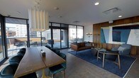 This contemporary single-floor space is thoughtfully designed to serve as both a social gathering and collaborative lounge area. The open-plan layout seamlessly blends a dining/work zone with a relaxed seating area, creating a versatile and welcoming environment.

The dining and work area features a substantial wooden communal table paired with plush teal velvet chairs, suspended above by an elegant curtain-style pendant light fixture that adds a distinctive architectural touch to the ceiling.

Adjacent to the dining zone, the lounge area is anchored by a rich cognac leather sofa set atop a deep navy blue area rug, complemented by a sculptural floor lamp and a warm wood-paneled accent wall. A teal abstract artwork introduces a bold pop of color, adding visual interest to the space.

Large floor-to-ceiling windows and glass sliding doors flood the interior with natural light while offering clear sightlines to an expansive outdoor terrace. The terrace, furnished with seating and tables, extends the usable space and provides elevated urban views of the surrounding cityscape.

The overall ambiance is modern and refined, combining warm wood tones, rich jewel-toned textiles, and sleek dark metal accents against a neutral palette of light grey walls and concrete-style flooring.