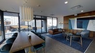 This contemporary single-floor space is thoughtfully designed to serve as both a social gathering and collaborative lounge area. The open-plan layout seamlessly blends a dining/work zone with a relaxed seating area, creating a versatile and welcoming environment.

The dining and work area features a substantial wooden communal table paired with plush teal velvet chairs, suspended above by an elegant curtain-style pendant light fixture that adds a distinctive architectural touch to the ceiling.

Adjacent to the dining zone, the lounge area is anchored by a rich cognac leather sofa set atop a deep navy blue area rug, complemented by a sculptural floor lamp and a warm wood-paneled accent wall. A teal abstract artwork introduces a bold pop of color, adding visual interest to the space.

Large floor-to-ceiling windows and glass sliding doors flood the interior with natural light while offering clear sightlines to an expansive outdoor terrace. The terrace, furnished with seating and tables, extends the usable space and provides elevated urban views of the surrounding cityscape.

The overall ambiance is modern and refined, combining warm wood tones, rich jewel-toned textiles, and sleek dark metal accents against a neutral palette of light grey walls and concrete-style flooring.