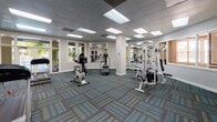 Located at 1060 Coral Ridge Dr in Coral Springs, Florida, this fitness center offers a well-equipped, single-floor exercise facility designed to support a well-rounded workout routine. The space features a drop-panel ceiling with recessed fluorescent lighting that ensures consistent, bright illumination throughout. The floor is covered in a distinctive patterned carpet in teal, blue, and green tones, adding a modern, energetic feel to the environment.

The main workout area is generously sized, providing ample room for a variety of cardio and strength training equipment, including stationary bikes, an elliptical, a stair climber, treadmills, and a multi-station cable weight machine. Large mirrors line sections of the walls, enhancing the sense of space and allowing users to monitor their form during exercise.

Natural light flows into the facility through multiple windows and glass-paneled doors, with views of lush tropical landscaping and direct visual access to an outdoor pool and patio area. A wall-mounted television is positioned near the entrance, offering entertainment during workouts.

The entryway features a vibrant lime-green accent wall that adds a pop of color and energy to the space. Informational boards and a sanitizing station are conveniently located near the entry. The overall ambiance is clean, functional, and inviting, reflecting a commitment to resident wellness in a bright Florida setting.