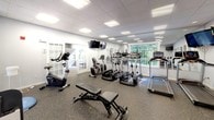 This fitness center is a comprehensively outfitted facility designed to support a full range of workout routines. The space features a drop-panel ceiling with recessed and flush lighting, light-toned walls, and durable rubber flooring throughout, creating a clean and functional environment. Large windows allow generous natural light to filter in, offering views of the surrounding greenery and contributing to an open, airy atmosphere. Full-length mirrors along the walls further enhance the sense of space.

The cardio section is well-stocked with multiple treadmills, elliptical trainers, a recumbent bike, an upright stationary bike, and a rowing machine, providing ample options for cardiovascular training. Wall-mounted flat-screen televisions are positioned throughout for entertainment during workouts.

The strength training area features a multi-station cable machine by Body-Solid, a dumbbell rack, adjustable weight benches, and additional resistance equipment. Functional fitness accessories including a BOSU balance trainer, stability balls, and exercise mats round out the offerings for flexibility and balance training.

French-style glass doors connect sections of the facility, adding an elegant architectural touch. A posted rules board and informational signage reflect an organized, community-oriented environment. Overall, this fitness center is thoughtfully arranged to accommodate a diverse range of fitness goals.