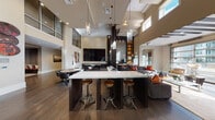 This contemporary amenity space is designed to impress, combining sophisticated design with a wide range of social and recreational features across a single expansive floor. The main gathering area commands attention with dramatically soaring double-height ceilings lined with clerestory windows that flood the space with natural light. Dark hardwood flooring anchors the open-plan layout, while a central bar-height counter with a crisp white surface and industrial-style leather barstools serves as a natural focal point. Pendant light fixtures with crystal-like glass casings hang overhead, adding elegance to the room.

A dedicated billiards area features a sleek black pool table, wall-mounted cue storage, bold artwork, and a large reflective wall display, creating an energetic game room atmosphere. Adjacent to this is a kitchenette outfitted with dark countertops, a stainless steel sink, microwave, and a standout geometric-patterned metallic tile backsplash that extends dramatically up the wall.

A separate media lounge offers a more intimate setting, with plush seating, soft carpeting, and a floor-to-ceiling custom shelving wall housing a large-screen television. Throughout the space, curated décor elements — including a striking wall installation of natural wood cross-sections and vibrant artwork — lend the space a warm, artistic character that balances modern design with organic texture.
