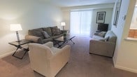 This single-floor space offers a warm and functional living environment designed for everyday comfort. The living room serves as the heart of the space, showcasing a generous, open layout with soft wall-to-wall carpeting that adds a cozy feel underfoot. A large sliding glass door with vertical blinds floods the room with natural light, creating a bright and airy atmosphere while providing access to the outdoors. The seating arrangement features a full sofa with accent pillows, a plush armchair, and a loveseat, all centered around a dark-toned coffee table — ideal for relaxed gatherings or quiet evenings at home. A floor lamp provides additional ambient lighting, while a flat-screen television and framed wall art contribute to the room's lived-in, homey character. The neutral color palette of soft greens and beiges throughout the walls and furnishings creates a calm, cohesive aesthetic that feels both inviting and versatile. The layout is thoughtfully arranged to maximize usable space while maintaining an open, uncluttered feel. Overall, this space combines practical design with comfortable living, making it a well-suited environment for daily life.
