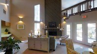 This inviting home showcases impressive architectural design centered around a striking floor-to-ceiling wood-paneled fireplace wall that serves as the dramatic focal point of the main living area. The soaring ceilings create an expansive sense of volume, enhanced by tall vertical windows that flood the space with abundant natural light and offer views of the surrounding trees. A loft overlook with a classic wooden railing adds vertical dimension and architectural interest to the open layout. The living area is thoughtfully arranged with comfortable seating, including plush armchairs and a sofa, complemented by warm accent lighting that creates an inviting ambiance throughout. A neutral color palette of soft taupes and creams runs throughout the space, accented by touches of yellow and natural wood tones that add warmth and character. A distressed white sideboard serves as a functional and decorative centerpiece, while candlestick accents and carefully selected décor complete the refined, transitional aesthetic. The open floor plan and seamless flow between spaces make this home both practical and visually engaging, offering a welcoming atmosphere suited to both everyday living and entertaining.