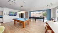 Located at 100 Seventh Street in Pittsburgh, PA, this single-floor community space is a lively and thoughtfully designed lounge offering a wide range of amenities. The open-plan layout is flooded with natural light, courtesy of expansive floor-to-ceiling windows and sliding glass doors that open directly onto an outdoor terrace with urban views.

The living area serves as the heart of the space, featuring a curved teal sectional sofa accented with vibrant green and patterned throw pillows, a colorful plaid area rug, and a large wall-mounted flat-screen TV set into a bold blue accent niche flanked by built-in shelving. Warm hardwood-style flooring runs throughout, creating a cohesive and inviting atmosphere.

A dedicated games zone adds recreational appeal, housing a full-size pool table and a shuffleboard table, both positioned to take advantage of the natural light streaming through the large windows. A statement pendant light overhead and decorative wall art enhance the character of this area.

The kitchen is clean and contemporary, showcasing light wood cabinetry with brass hardware, sleek countertops, a stainless steel double sink, and a striking turquoise tile backsplash that adds a pop of color. A dining table with mid-century modern chairs provides a comfortable space for gathering and collaboration. The overall ambiance is bright, energetic, and welcoming.