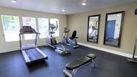 This single-floor fitness room offers a clean, functional space designed for effective workouts. The room is finished with neutral beige walls and durable dark rubber flooring, providing a practical surface well-suited for exercise equipment. Natural light fills the space through multiple white-framed windows fitted with blinds, offering views of surrounding greenery while keeping the atmosphere bright and welcoming. Recessed ceiling lights supplement the natural light, ensuring consistent illumination throughout. The room is equipped with a treadmill, an elliptical trainer, a recumbent stationary bike, and a workout bench, covering a solid range of cardiovascular and strength training options. Two large, black-framed mirrors are mounted on the wall opposite the windows, enhancing the sense of space and allowing users to monitor their form during exercise. The layout is thoughtfully arranged to allow comfortable movement between machines. White baseboards provide a crisp contrast to the darker flooring, contributing to the room's clean, well-maintained appearance. Overall, the space delivers a straightforward and practical fitness environment.