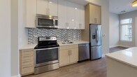 Located at 150 River's Edge Dr in Medford, MA, this single-floor residence offers a thoughtfully designed, modern living space with clean lines and contemporary finishes throughout. The open-concept layout connects the living area to a well-equipped kitchen, which features stainless steel appliances including a range, over-the-range microwave, dishwasher, and French door refrigerator. Two-tone cabinetry — warm wood lower cabinets paired with crisp white upper cabinets — frames a mosaic tile backsplash and light-colored countertops, creating a stylish and functional cooking space. A peninsula with pendant lighting provides additional prep space and serves as a natural divider between the kitchen and the open living area. Warm-toned hardwood-style flooring flows consistently throughout the main living spaces, enhancing the sense of continuity and warmth. The bedroom offers a generous walk-in closet with built-in shelving, providing ample storage. The bathroom is finished with large-format tile flooring, a white vanity with granite-style countertop, an integrated sink, and a well-lit mirror above. Neutral gray walls and white trim contribute to the space's bright, modern ambiance. The entry features a secure keypad door lock, adding a contemporary touch to everyday convenience.