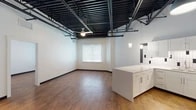 This single-floor unit showcases a striking industrial-modern aesthetic defined by its most prominent architectural feature: an exposed black steel ceiling grid with visible structural beams, ductwork, and piping running throughout the open living areas. Warm hardwood-style flooring and crisp white walls provide a clean contrast to the raw overhead elements, creating a balanced and visually engaging interior.

The open-concept living area is generously proportioned, with a ceiling fan and natural light from a window fitted with white blinds. This space flows seamlessly into the kitchen, which is outfitted with white shaker-style cabinetry extending to the upper walls, a white quartz countertop, a central island, and a full suite of stainless steel appliances including a refrigerator, range, microwave, and dishwasher. A white subway tile backsplash with black accent tiles adds a modern decorative touch.

The bedroom areas share the same hardwood flooring and soaring exposed ceilings, each with ceiling fans and windows for natural light. A spacious walk-in closet with wire shelving and hanging rods provides ample storage. The bathroom features a frameless glass-enclosed walk-in shower with floor-to-ceiling tile surround, complemented by white and black accent tile wainscoting.