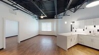 This single-floor unit showcases a striking industrial-modern aesthetic defined by its most prominent architectural feature: an exposed black steel ceiling grid with visible structural beams, ductwork, and piping running throughout the open living areas. Warm hardwood-style flooring and crisp white walls provide a clean contrast to the raw overhead elements, creating a balanced and visually engaging interior.

The open-concept living area is generously proportioned, with a ceiling fan and natural light from a window fitted with white blinds. This space flows seamlessly into the kitchen, which is outfitted with white shaker-style cabinetry extending to the upper walls, a white quartz countertop, a central island, and a full suite of stainless steel appliances including a refrigerator, range, microwave, and dishwasher. A white subway tile backsplash with black accent tiles adds a modern decorative touch.

The bedroom areas share the same hardwood flooring and soaring exposed ceilings, each with ceiling fans and windows for natural light. A spacious walk-in closet with wire shelving and hanging rods provides ample storage. The bathroom features a frameless glass-enclosed walk-in shower with floor-to-ceiling tile surround, complemented by white and black accent tile wainscoting.