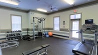 This single-floor fitness room offers a practical and well-equipped space for residents or members seeking a convenient workout facility. The room features rubber flooring throughout, providing a durable and safe surface ideal for exercise. A multi-station cable machine serves as the centerpiece for strength training, complemented by a weight bench and an assortment of free weights and accessories. Stability balls in multiple sizes are neatly stored along the wall, supporting flexibility and core training options.

The walls are finished in a light neutral tone accented by horizontal striped trim in muted blue and tan, lending a clean, structured aesthetic to the space. A ceiling fan overhead aids in air circulation during workouts, while fluorescent lighting ensures consistent illumination throughout. Natural light filters into the room through two windows fitted with blinds, as well as a glass-paneled entry door that enhances the bright, open feel. An emergency exit sign is visible, reflecting proper safety standards. The overall layout is efficient and organized, making effective use of the available floor space to accommodate a variety of exercise routines.
