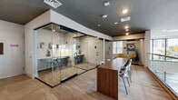 Located at 435 Main St in Hackensack, NJ, this contemporary single-floor commercial space is designed to foster productivity and collaboration. The layout features a dynamic mezzanine-level upper floor with a sleek glass railing and warm wood-capped balustrade that overlooks a bright, open lower-level lounge area below, where large street-facing windows flood the space with natural light and views of the surrounding neighborhood.

The upper level hosts a central long communal work table in rich walnut-toned wood, accompanied by modern mixed-pattern seating. Glass-enclosed private office rooms with black-framed sliding doors line one side, offering acoustic separation while maintaining visual openness throughout. Each enclosed room is equipped with built-in desks and task seating, ideal for focused individual or small group work.

Accent walls finished in textured concrete and warm earth tones provide an industrial character balanced by clean white walls and recessed lighting overhead. A dedicated workstation nook with built-in desk and wall-mounted lighting adds functional variety. The lower level features an inviting lounge furnished with accent chairs and lush greenery. Light wood-plank flooring runs throughout, unifying the space with a warm, professional ambiance well-suited for a modern office or coworking environment.