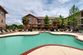 Located at 200 Long St in Slidell, Louisiana, this outdoor amenity space serves as a central gathering area for the surrounding multi-story residential community. At the heart of the space is a generously sized freeform swimming pool with a distinctive curved, organic shape and a classic brick-trimmed border. The pool deck is expansive, offering ample room for lounging, and is outfitted with numerous sling-style lounge chairs and patio seating arranged around the perimeter for residents to relax and socialize. A wooden pergola structure anchors one side of the deck, providing a shaded outdoor gathering spot beneath its open-beam canopy. Mature palm trees and lush greenery are strategically placed throughout the area, lending a tropical, resort-like ambiance well-suited to the Louisiana climate. The space is enclosed by a decorative iron fence with a secure gated entry, ensuring a private and controlled environment. Manicured landscaping with shrubs and ground cover adds visual appeal along the fence line. The surrounding multi-story residential buildings, finished in warm earth-toned stucco, frame the courtyard on multiple sides, with private balconies overlooking the pool area. The overall atmosphere is open, bright, and inviting, ideal for outdoor recreation and leisure.