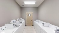 This shared laundry facility is a single-floor space designed for practical, efficient use. The room is arranged with two parallel rows of white top-load washing machines lining both the left and right walls, providing ample capacity for multiple users. Each machine is neatly positioned and appears well-maintained, contributing to the room's orderly atmosphere.

The interior features neutral gray walls that create a clean, modern aesthetic, while light-toned tile flooring adds brightness and is easy to maintain. Overhead, a rectangular flush-mount fluorescent light fixture provides strong, even illumination throughout the space, ensuring excellent visibility.

At the far end of the room, a light wood-tone door with a small window insert serves as the primary entry or exit point, allowing a modest amount of additional light while maintaining privacy. A ventilation grate is visible in the ceiling, supporting proper air circulation — an important feature in a laundry environment.

Overall, the space is straightforward and functional, prioritizing accessibility and cleanliness. Its simple, uncluttered design makes it easy to navigate and maintain, reflecting a well-planned utility room built to serve the needs of multiple users efficiently.