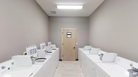 This shared laundry facility is a single-floor space designed for practical, efficient use. The room is arranged with two parallel rows of white top-load washing machines lining both the left and right walls, providing ample capacity for multiple users. Each machine is neatly positioned and appears well-maintained, contributing to the room's orderly atmosphere.

The interior features neutral gray walls that create a clean, modern aesthetic, while light-toned tile flooring adds brightness and is easy to maintain. Overhead, a rectangular flush-mount fluorescent light fixture provides strong, even illumination throughout the space, ensuring excellent visibility.

At the far end of the room, a light wood-tone door with a small window insert serves as the primary entry or exit point, allowing a modest amount of additional light while maintaining privacy. A ventilation grate is visible in the ceiling, supporting proper air circulation — an important feature in a laundry environment.

Overall, the space is straightforward and functional, prioritizing accessibility and cleanliness. Its simple, uncluttered design makes it easy to navigate and maintain, reflecting a well-planned utility room built to serve the needs of multiple users efficiently.