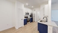 Located at 5333 E Parmer Lane in Austin, TX, this one-bedroom, one-bathroom unit offers a thoughtfully designed single-floor layout that maximizes every square foot. Light wood-style flooring flows continuously throughout the space, complementing the crisp white walls and recessed lighting that keep the interior feeling bright and open.

The kitchen serves as a visual centerpiece, showcasing bold navy blue lower cabinetry paired with white upper cabinets and speckled granite-style countertops. Stainless steel appliances — including a range, microwave, and refrigerator — provide a sleek, modern finish. A kitchen island with a stainless steel sink adds both functionality and style.

The open living area benefits from a large window dressed with horizontal blinds, allowing natural light to fill the room while offering views of the surrounding greenery. The space connects seamlessly to the entry, which features clean white walls and a hallway leading to the bedroom, which includes a spacious double-door closet.

The bathroom continues the navy and granite design language, featuring a wide vanity with an under-mount sink, a large frameless mirror, and stylish pendant lighting — creating a polished, cohesive aesthetic that ties the entire unit together.
