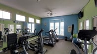 This fitness center offers a practical and energizing environment designed to support a full cardio and exercise routine. The space is finished with a two-tone color scheme — bold teal and lime green accent walls — that lends a vibrant, motivating atmosphere throughout. A ceiling fan centered overhead provides air circulation, complementing the abundant natural light that pours in through multiple windows fitted with white plantation shutters along two walls. The flooring is a durable, neutral-toned carpet suited for a high-traffic workout environment. The cardio area is well-stocked with multiple treadmills, an elliptical trainer, and a stair-climber machine, all arranged to maximize the available floor space. A wall-mounted mirror on one side visually expands the room and allows users to monitor their form during exercise. Exercise balls in multiple sizes are stored in a corner alongside a small equipment station. Informational postings and safety notices are displayed on the walls, reinforcing the space's functional purpose. The overall layout is thoughtfully organized, creating an open, accessible fitness facility that is both practical and welcoming for everyday use.
