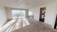 Located at 2101 SW Sunset Blvd in Renton, Washington, this single-floor unit offers a well-organized layout designed for comfortable everyday living. The open living area is generously sized, featuring plush carpeting, a textured ceiling, and large sliding glass doors flanked by vertical blinds that allow abundant natural light to pour in and provide direct access to an outdoor view of surrounding trees. A secondary window further brightens the space. The living area flows naturally into the kitchen, which is equipped with white shaker-style cabinetry, dark countertops, a white electric range with overhead ventilation, and a stainless steel sink — all set atop wood-look flooring for easy maintenance. The unit includes multiple carpeted bedrooms, each featuring sliding windows with baseboard heating beneath, offering a cozy and functional retreat. The bathroom is appointed with a full soaking tub and shower combination, accented by clean white surround panels and wood-look flooring. Neutral wall tones and textured ceilings are consistent throughout, lending the space a cohesive, understated aesthetic. A dedicated entry area with a closet completes the layout, adding practical storage to this well-rounded ground-floor residence.