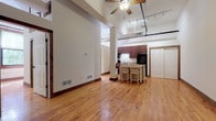 This single-floor loft-style space showcases a compelling blend of industrial heritage and contemporary living. Throughout, warm-toned hardwood floors extend seamlessly, complemented by rich wood baseboards and trim that add a refined contrast to the open layout.

The main living area is anchored by soaring ceilings fitted with a ceiling fan and track lighting, creating a bright and airy atmosphere. A structural column punctuates the open floor plan, adding to its industrial character. The kitchen is well-appointed with rich dark wood cabinetry, granite countertops, black appliances including a range, refrigerator, and dishwasher, and a freestanding island with a stainless steel top. Large sliding doors provide convenient storage access nearby.

Several versatile rooms branch off from the main area, each featuring gleaming hardwood floors. One room boasts exposed white-painted brick walls and tall windows that invite abundant natural light. Another room with exposed brick features a charming barn-style sliding door. The entry area displays distinctive reclaimed hardwood flooring, a bold red interior door with wood trim, and an electrical panel.

Two bathrooms serve the space — one full bath with a soaking tub, dark wood vanity, and wood-trimmed mirror, and a second bath showcasing exposed white brick walls. The overall ambiance is one of urban loft character with warm, residential touches throughout.