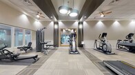 Located on Polo Club Drive in Strongsville, Ohio, this fitness center offers a comprehensive and thoughtfully designed workout facility spread across a single open floor. The space features a modern aesthetic with neutral-toned walls, recessed lighting, and a combination of pendant light fixtures and ceiling fans that contribute to a comfortable and well-lit environment. The layout is generously proportioned, providing ample room to move freely between equipment zones. The cardio section is prominently featured and includes multiple Matrix treadmills, elliptical trainers, a stair climber, and a rowing machine, each positioned to allow comfortable spacing between users. Flat-screen televisions are mounted throughout, adding an entertainment element to cardio sessions. A dedicated strength training area houses a multi-function cable machine along with benches and free weight equipment. Large framed mirrors are strategically placed to enhance the sense of space and allow users to monitor their form. Interior glass partitions and windows allow natural light to filter into portions of the space, creating an open, airy feel. Patterned carpet tiles cover the floor throughout, providing both functionality and a polished visual appeal. The overall ambiance is clean, professional, and well-maintained, making this fitness center a practical and inviting space for a wide range of exercise routines.