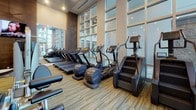 This Health & Wellness Center is a thoughtfully designed, single-floor fitness facility offering a comprehensive range of equipment and dedicated activity zones. The main gym area features striped, multi-tone carpeting and soaring ceilings with recessed lighting, creating a bright and energizing atmosphere. Floor-to-ceiling glass curtain walls line key sections of the space, allowing abundant natural light and outdoor views to enhance the workout environment.

The cardio zone is lined with treadmills, ellipticals, and stair climbers positioned along the expansive window wall, offering views while exercising. The strength training area is well-stocked with multi-tiered dumbbell racks, cable machines, a functional training rig, and resistance equipment, all reflected in mirrored walls that amplify the sense of space.

A dedicated studio room features a herringbone hardwood floor, ceiling fans, wall-mounted TVs, full-length mirrors, and spin bikes, making it ideal for cycling and group fitness classes. This studio also provides stability balls, yoga mats, and hand weights for varied workouts.

A built-in shelving unit with a compact beverage refrigerator serves as a convenient amenity station. The facility's overall design balances functionality with a polished, modern aesthetic.