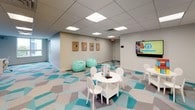 This thoughtfully designed play room offers a bright, engaging environment tailored for children. The space is unified by a striking geometric-patterned carpet in teal, gray, and white tones that sets a playful foundation throughout. Overhead, a drop-ceiling with recessed panel lighting ensures consistent, even illumination across the entire room.

The main activity area features multiple child-sized round white tables paired with matching chairs, providing ample space for group play and creative activities. Soft teal bean bag chairs are scattered throughout, offering casual seating options for relaxed play or media viewing. A wall-mounted flat-screen television serves as a focal point on one end of the room, ideal for educational or entertainment programming.

Along one wall, a built-in white cabinetry unit with upper cabinets and a small sink provides convenient storage and a functional workspace for supervisors or caregivers. Adjacent open shelving units are stocked with a variety of toys, games, and building blocks, keeping activities within easy reach. A dedicated desk area with a task chair is tucked into a corner accented by a bold teal feature wall, offering a quieter space for focused activities.

Decorative framed art panels line the walls, and a window in the corner nook brings in natural light, completing this well-organized and inviting play environment.