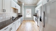 Located at 1193 Waxwing Dr, Building 1185 Unit B in Florence, SC, this two-floor residence combines classic architectural details with modern finishes throughout. The main living and dining areas are defined by striking embossed tin ceilings, elegant arched openings, and white decorative columns that add a refined, traditional character. Warm wood-look flooring flows through the open-plan living spaces, complemented by soft sage-green and neutral beige wall tones. Ceiling fans are present throughout, ensuring comfort in every room. The galley-style kitchen is well-equipped with stainless steel appliances, including a range, microwave, and refrigerator, paired with crisp white cabinetry, dark granite countertops, and a classic subway tile backsplash. A window above the sink allows natural light to fill the space. The primary bedroom features a tray ceiling with crown molding, adding an elevated sense of space, while additional bedrooms are finished with neutral tones and tile flooring. Bathrooms are thoughtfully appointed with granite-topped white vanities, undermount sinks, and soaking tubs with tile surrounds. The upper level is accessed via a staircase visible from the main living area, offering additional living space. Overall, the home presents a cohesive blend of decorative craftsmanship and practical design.