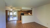 This single-story space showcases an open-concept design that connects the living area and kitchen seamlessly. The kitchen is defined by a raised peninsula counter with a light granite-style surface, providing both workspace and a natural divider between the cooking and living areas. Rich wood cabinetry lines the kitchen walls, offering generous storage, while a black refrigerator and built-in range hood are integrated into the layout. A recessed soffit detail above the kitchen adds architectural interest to the ceiling plane. The textured popcorn ceiling is present throughout, and a ceiling fan is visible in the adjacent living area, enhancing air circulation. Warm-toned hardwood flooring runs continuously across the main living space, adding cohesion and a sense of warmth. Mirrored sliding closet doors near the living area reflect natural light, contributing to a brighter interior. Sliding glass doors at the far end of the space allow abundant natural light to flow in and provide a connection to an outdoor area. The overall layout is practical and functional, with a straightforward flow between spaces.
